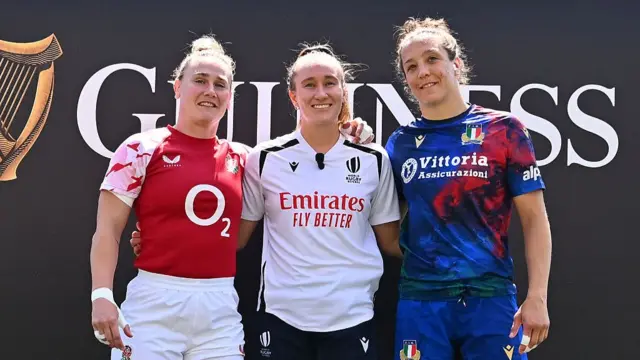 England captain Meg Jones (left, wearing a red training shirt and white shorts) and Italy captain Elisa Giordano (right, blue and red top with blue shorts) pose with their arms around the referee