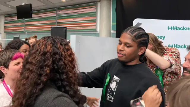 A young man at an election count inside a modern civic building hugs a supporter
