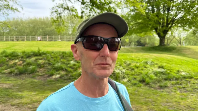 The image shows a man wearing a hat, sunglasses and a bright blue t shirt. He's stood in a green space.