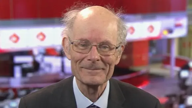 A man with grey, balding hair with wire-rimmed glasses wearing a suit, with the BBC studio behind him