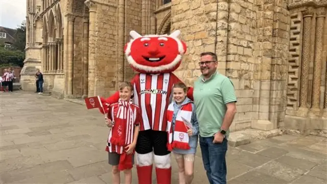 The club's imp mascot poses with the two children who wear scarves and football shirts and flags. Their father stands at the side of them.