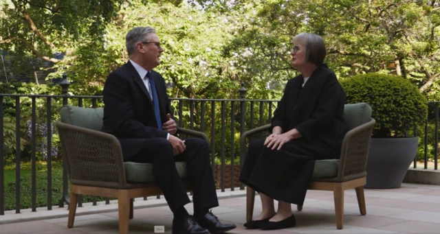 Prime Minister Keir Starmer (L) in a black suit sits down on a cushioned wooden chair, Baroness Harriet Harmen (L) sitting in a matching chair in a black dress. They're on a tiled deck in the back garden on 10 Downing Street