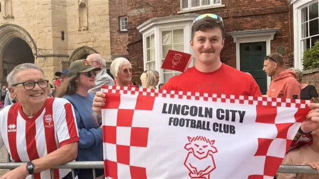 Aaron Trevor holds a Lincoln city FC flag out in front of him. Another fan smiles at him on his right.