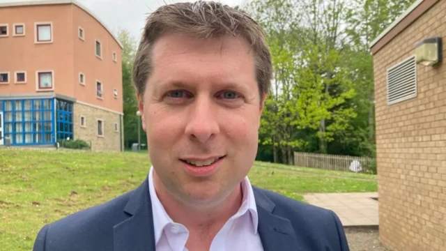 Richard John stands outside with a block of flats behind him. He has short brown hair and wears a blue suit.