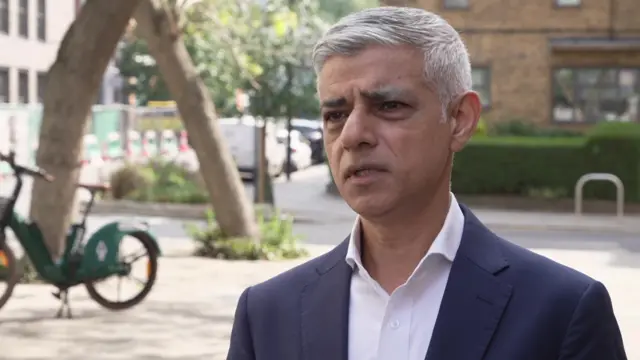 Mayor of London Sadiq Khan speaks to media wearing dark suit jacket and white shirt, with London street scene in background