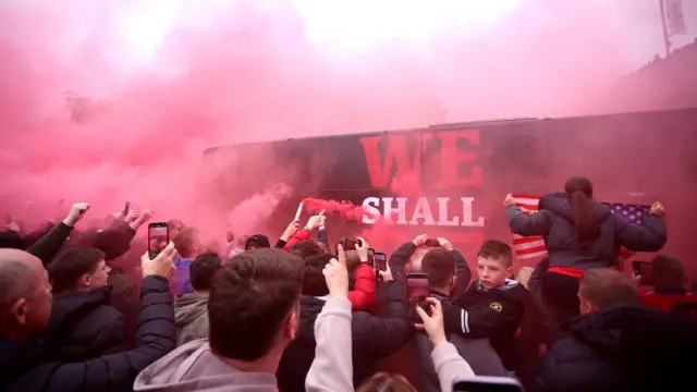 Fans and red smoke from flares can be seen billowing in front of the black Middlesbrough team bus which says "We Shall Be" in red and white writing on the side of it