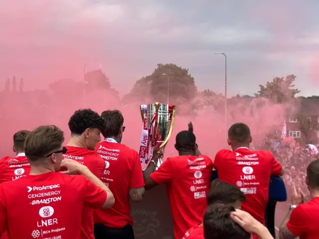 Red smoke fills the air and surrounds the players on the top of the bus.