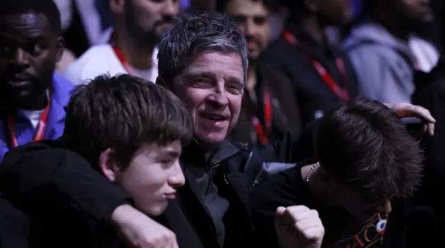 Noel Gallagher holds up his fist and has his arm around the shoulders of his child