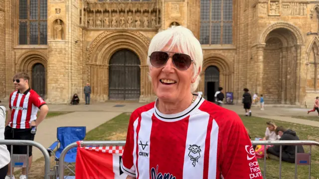 Andy stands in front of the Cathedral in his shirt. Other fans have started to gather in the background.