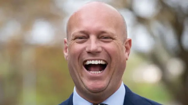 Dan Thomas laughs, he is wearing a navy suit and the background is blurred.