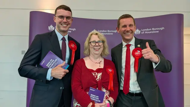 Three Labour councillors wearing red rosettes stand in front of a banner displaying the Hounslow Council logo.