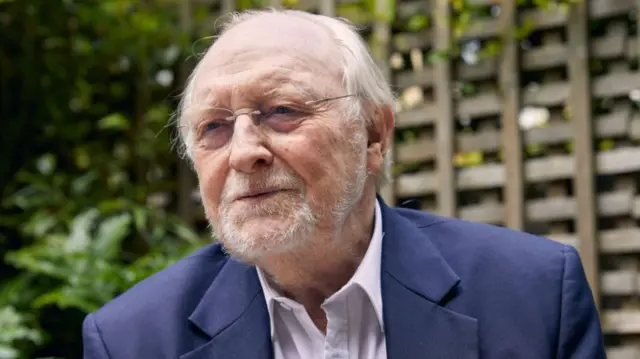 Neil Kinnock sits in a garden wearing a blue suit and glasses. He has balding white hair and a short white beard.