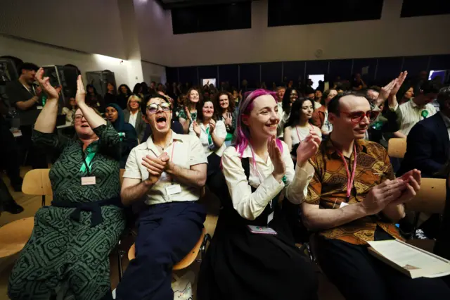 A group of activists, sitting in a packed room, cheer and applaud as results are announced.