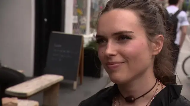 A woman stands on a pavement in London. She has her hair in a ponytail and wears a black top.
