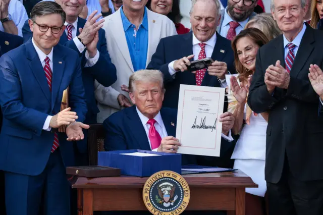 Trump sits at desk holding signed copy of bill, surrounded by clapping lawmakers