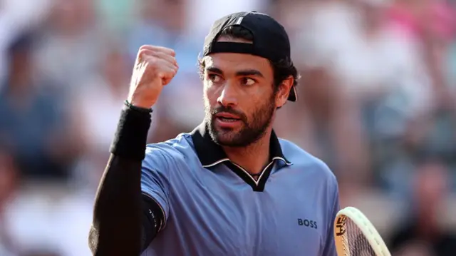 Matteo Berrettini, wearing a blue polo shirt with a black sleeve on his right arm, raises his right fist in celebration