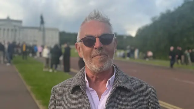 A man with short white hair is facing the camera wearing sunglasses