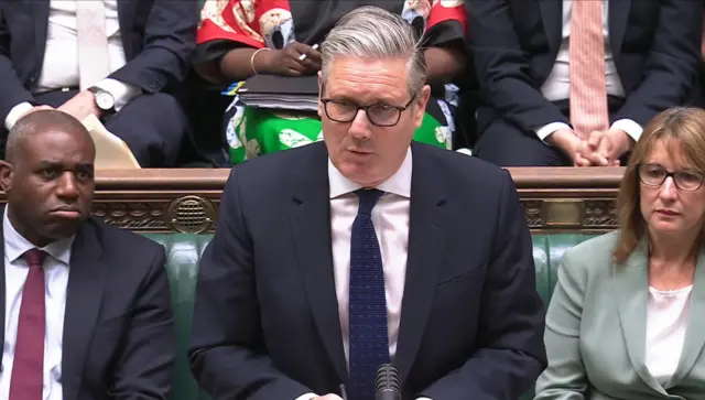 Keir Starmer standing at the dispatch box in the House of Commons during PMQs. Sitting behind him to his right is David Lammy, with Chancellor Rachel Reeves to his left