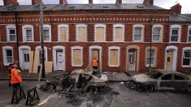 The husks of two burnt out cars are parked on the street in east Belfast in front of a row of terraced houses, some of which have their windows boarded up