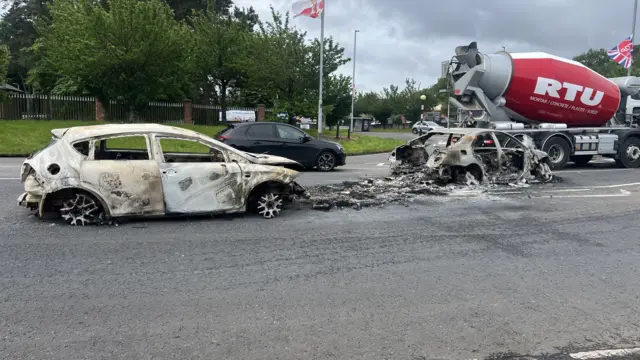 Two burnt out cars sit in the middle of a road as other traffic, including a large cement mixer truck, approach a roundabout