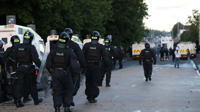 A number of police officers dressed in riot gear walk along a road alongside a number of police vehicles