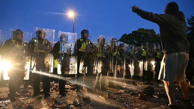A protester wearing shorts is standing at nighttime with his arms outstretched facing a line of riot officers with shields