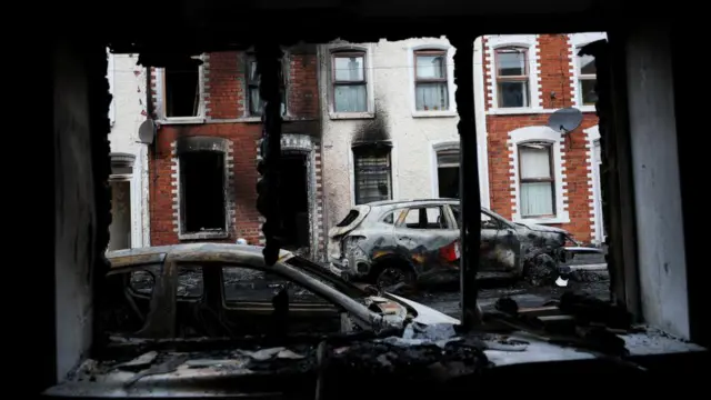 A view of two burnt cars as seen from the damaged window of a home.