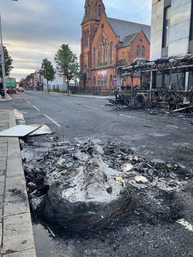 Charred remains of a burned out bus on the side of a road in east Belfast. The remaining structure of the Glider bus is to the left of the frame, on the opposite side of the road. In the background, the rest of the street, a red-brick church on the left side