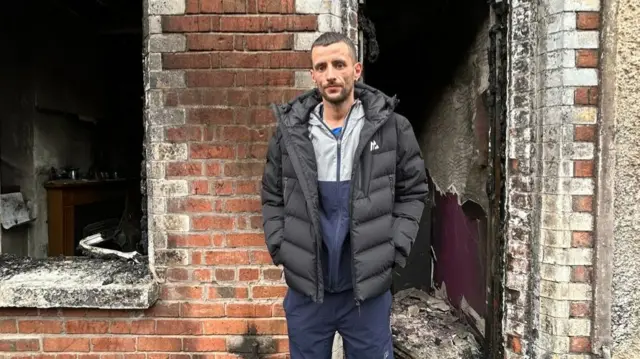 A man standing outside a burnt out red-brick house. He is wearing a navy and grey tracksuit and a black coat.