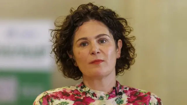 Claire Hanna wearing a flowery blouse. She has short, curly brown hair.