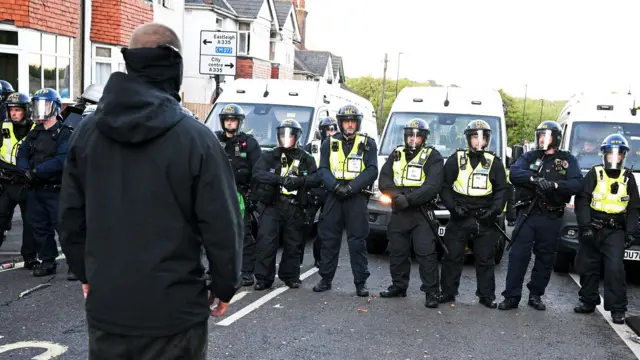 A man all dressed in black and wearing a black mask stands with his back to the camera, facing a row of police officers wearing protective face masks and helmets