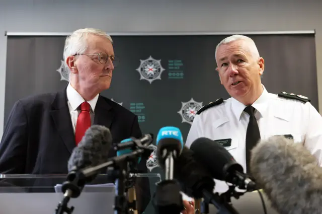 Hilary Benn (left) and Jon Boutcher (Right) adress the media in a room, several mics placed in front of them