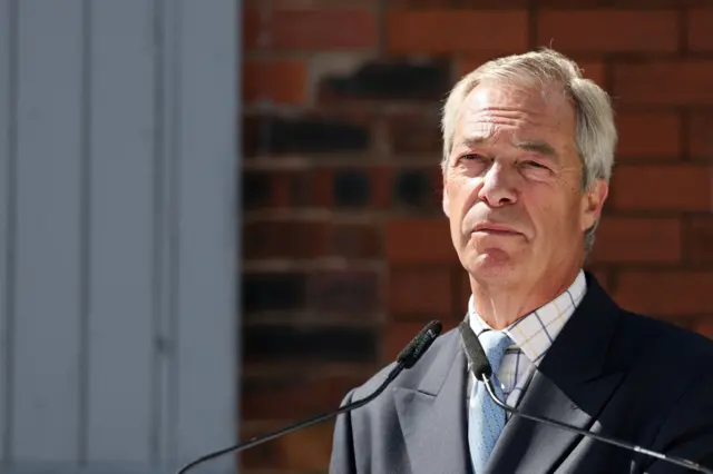 Nigel Farage in a dark suit and white checked shirt in front of some microphones