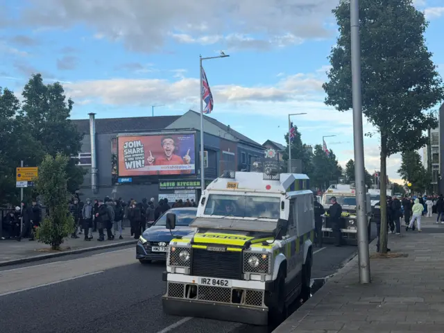 Photo of police land rover on road- behind it is a crowd in dark clothes
