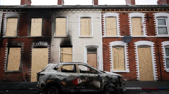 A burnt out car is in the fore of the image while residential properties have had boards placed where their windows were