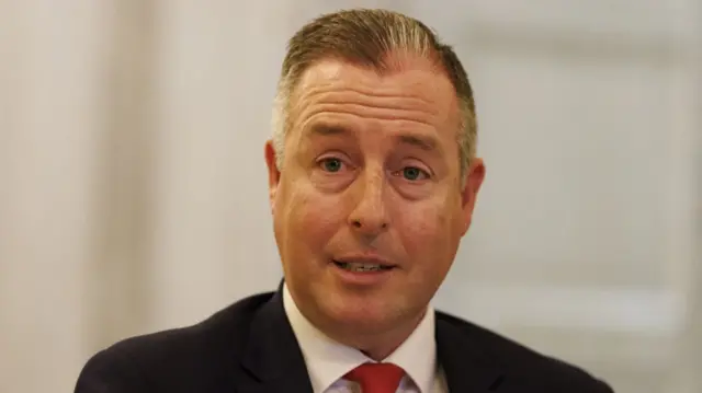 Paul Givan is wearing a black suit, red tie and white shirt