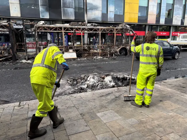 Men in high vis work gear brushing debris in front of the shell of a burnt out Glider bus in Belfast