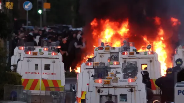 Police vehicles in the fore while a fire can be seen flaring just in front