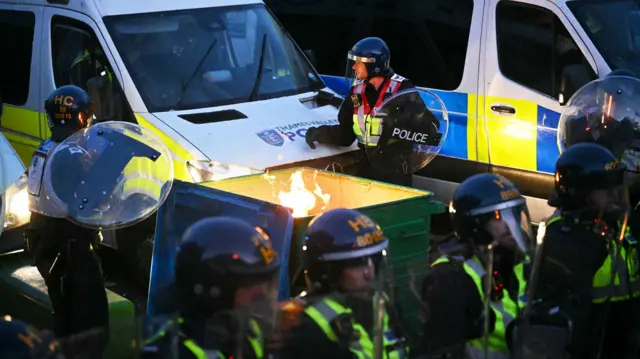 An officer wearing riot gear stands behind a commercial bin which is on fire. Behind him are a row of riot vans and in front of the bin are other officers wearing riot gear in a row