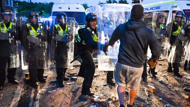 A police officer in riot gear pushes a shield towards a protester while other officers in riot gear look on