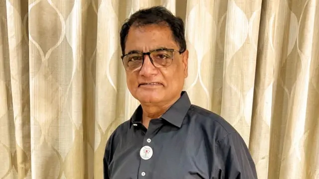 A man wearing glasses in a dark grey shirt standing in front of curtains.