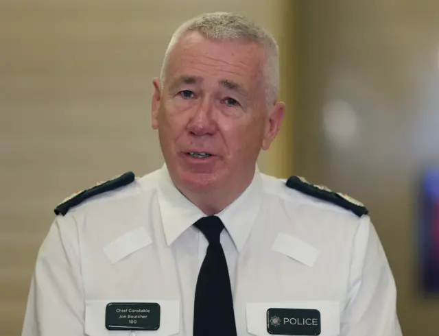 Jon Boutcher, a man with short white hair looks to the camera mid-speech during a press conference.  He is wearing a white shirt and a dark tie as part of his police uniform.