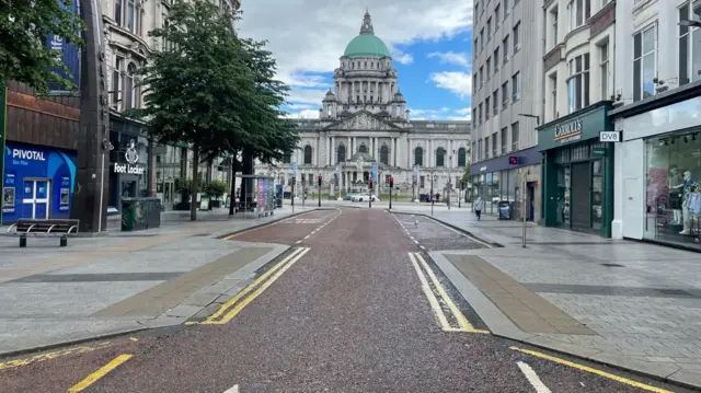 An empty Belfast city centre street, with no cars on the road and no people on the footpaths. The street is almost eerily quiet. On either side there are high street shops that appear to be closed. Facing the top of the street is a large building with a green dome, the city hall