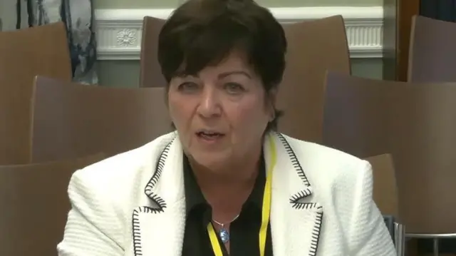 Rita Devlin has short black hair and is talking in the picture. She is wearing a black shirt and a white blazer. She has a yellow lanyard around her neck.