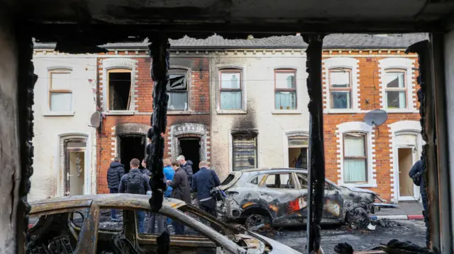 People stand next to burnt-out cars and homes