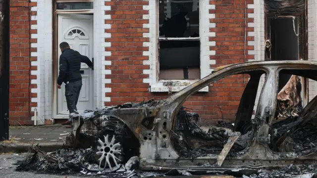 A burnt out car is seen on  Lendrick Street parked in front of a terraced home which has had its windows smashed and some of the front door burnt
