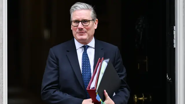 Keir Starmer, wearing a suit, walking out of 10 Downing Street with folders in his hands.