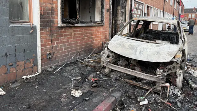 A burnt out car on the pavement, in front of a building which has had its windows smashed in.