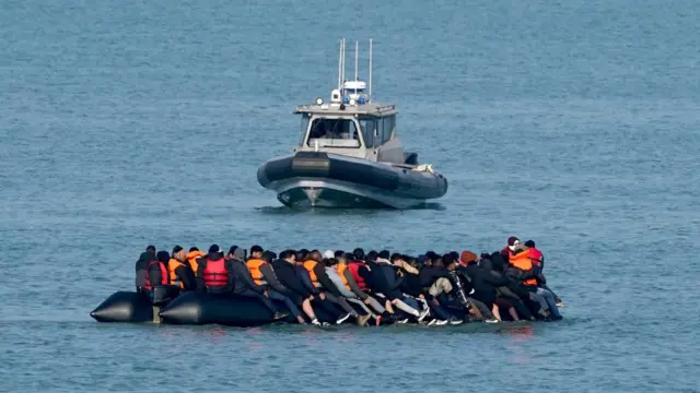 People on a dinghy in the water wearing life jackets. Behind them is a small vessel.