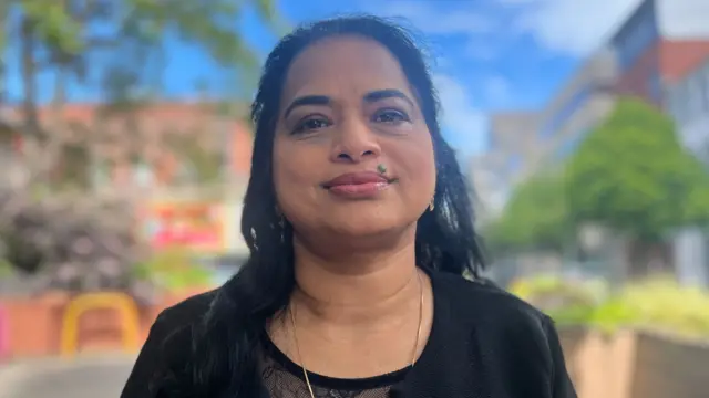 Biji Jose, a woman with long,straight back hair, looks to the camera on a city centre street. She is wearing a black jacket over a black and white top with lace detailing.